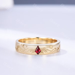 May include: A gold band ring featuring an engraved mountain range landscape with a crescent moon, stars, and a sun. A single kite-shaped red gemstone is set centrally within the band, positioned between the engraved mountain peaks.
