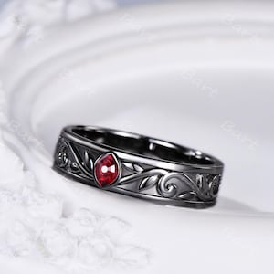 May include: A black metal ring featuring an intricate engraved leaf and vine pattern wrapping around the band. A single marquise-cut red gemstone is set prominently in the center of the design, contrasting against the dark, polished metal finish.