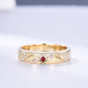 May include: A gold band ring featuring a celestial motif with a central red gemstone sun, two small round iridescent stones, two engraved planet shapes, and four small star-shaped accents set with clear white stones along the outer surface.