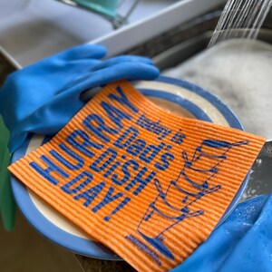 Op de afbeelding: Een oranje vaatdoek met blauwe tekst die "HURRAY today is dad's dish day!" luidt en een stapel blauw-wit servies.