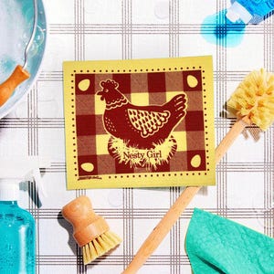 May include: A yellow dishcloth with a red and white checkered pattern and a brown illustration of a hen in a nest. The text "Nesty Girl" is printed below the hen.