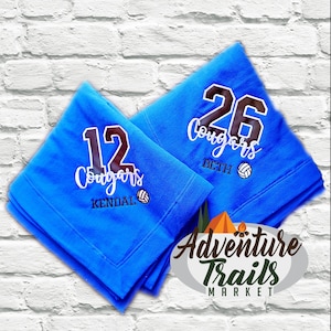 May include: Two blue fleece blankets with embroidered volleyball team names and numbers. The blankets are folded and stacked on top of each other. The top blanket has the number 26 and the name "Beth" embroidered on it. The bottom blanket has the number 12 and the name "Kendal" embroidered on it. Both blankets have the word "Cougars" embroidered on them. The blankets are from Adventure Trails Market.