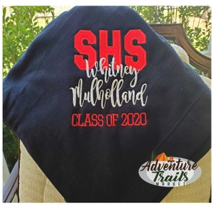 May include: A navy blue blanket with red and white embroidered text. The text includes "SHS", "Whitney Mulholland", and "CLASS OF 2020". The blanket is folded over a chair.