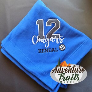 May include: Blue fleece blanket with the number 12, the word "Cougars", the name "KENDAL", and a volleyball graphic embroidered on it. The text "Adventure Trails Market" is also embroidered on the blanket.