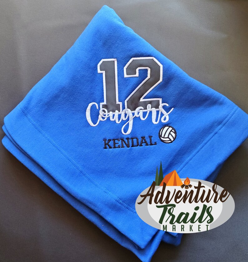 May include: Blue fleece blanket with the number 12, the word "Cougars", the name "KENDAL", and a volleyball graphic embroidered on it. The text "Adventure Trails Market" is also embroidered on the blanket.
