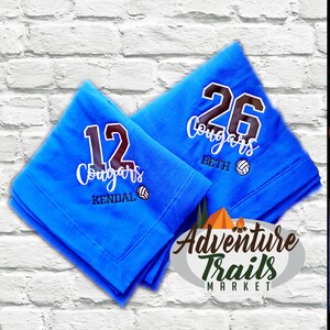 May include: Two blue fleece blankets with the word "Cougars" and a volleyball graphic. The blankets are personalized with the numbers "12" and "26" and the names "Kendal" and "Beth". The blankets are from Adventure Trails Market.