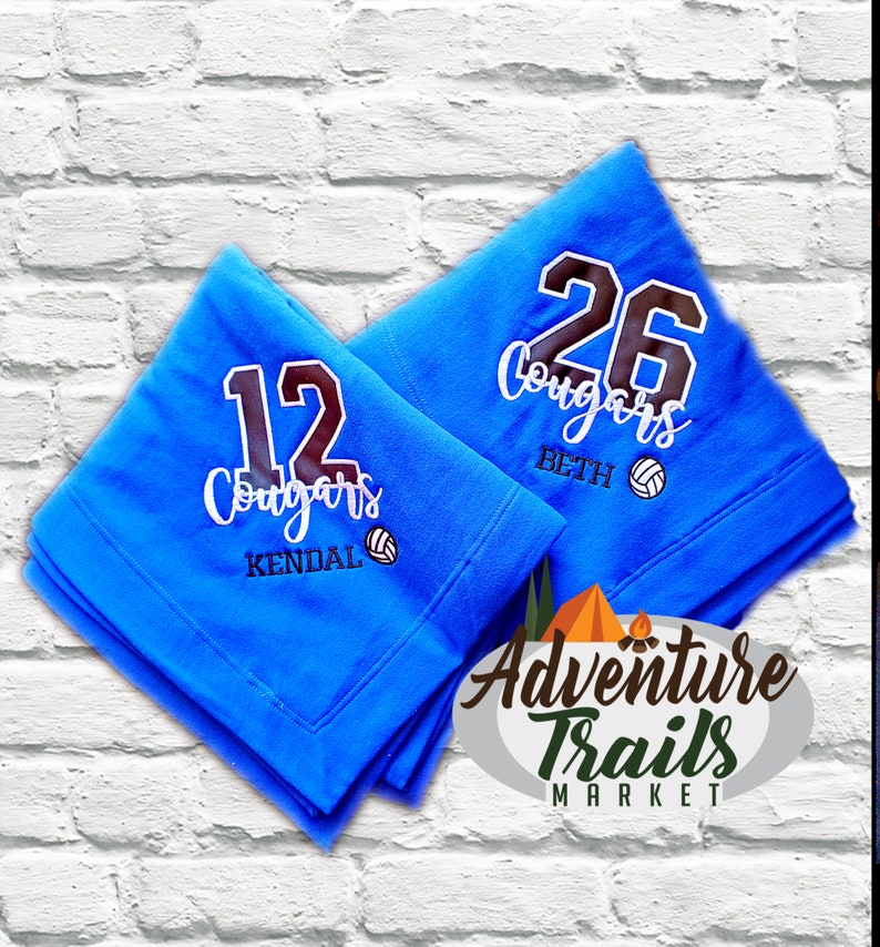 May include: Two blue fleece blankets with the word "Cougars" and a volleyball graphic. The blankets are personalized with the numbers "12" and "26" and the names "Kendal" and "Beth". The blankets are from Adventure Trails Market.