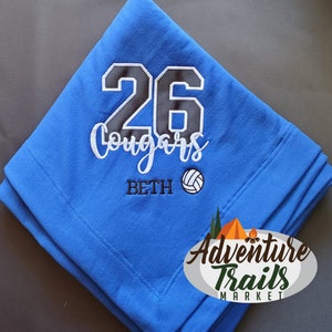 May include: Blue fleece blanket with white embroidery. The embroidery includes the number 26, the word "Cougars", the name "Beth", and a volleyball. The blanket is folded in half and the Adventure Trails Market logo is visible in the bottom right corner.
