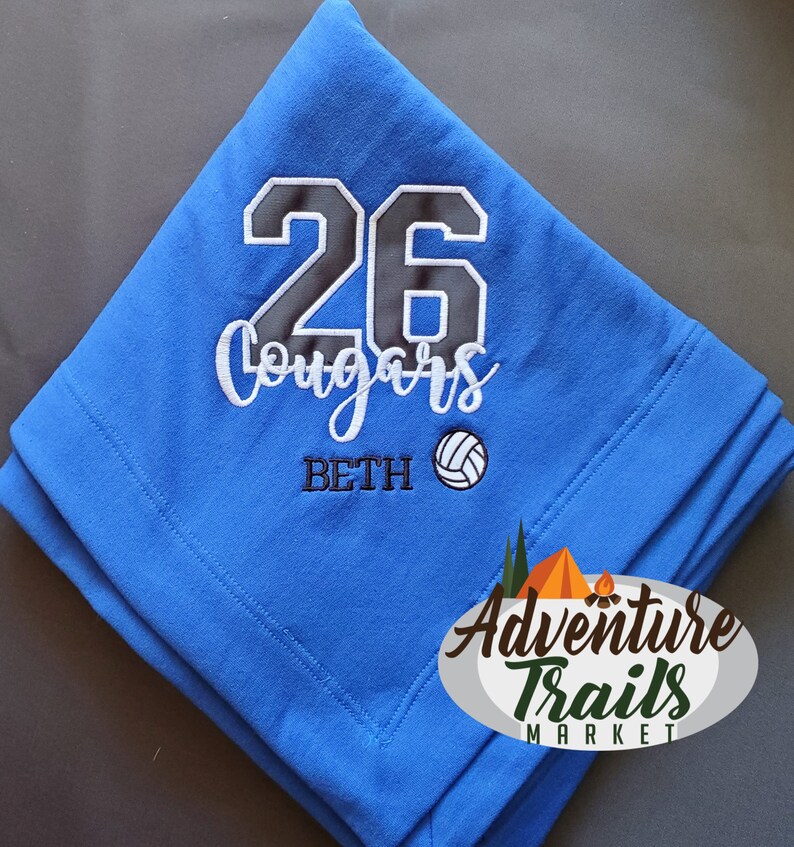 May include: Blue fleece blanket with white embroidery. The embroidery includes the number 26, the word "Cougars", the name "Beth", and a volleyball. The blanket is folded in half and the Adventure Trails Market logo is visible in the bottom right corner.