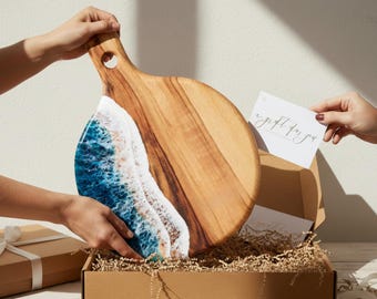 Peut inclure: Planche de service en bois avec une poignée, ornée d'un motif de vagues en résine bleue et blanche. La planche est tenue au-dessus d'une boîte en carton remplie de matériel d'emballage. Une carte cadeau est également incluse.