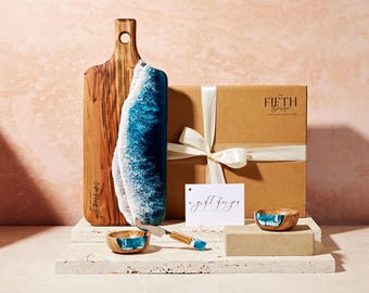 May include: A wooden serving board with a blue and white resin wave design. The board is packaged in a brown box with a white ribbon and a gift tag that says "a gift for you". The set also includes two small blue and white resin bowls and a butter knife.