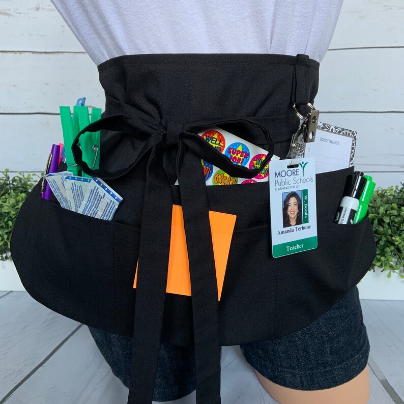 May include: Black teacher apron with multiple pockets, a black bow, and a name tag that reads "Moore Public Schools, Amanda Terhune, Teacher". The apron has a variety of stickers, including "Super Job", "Well Done", and "Great".