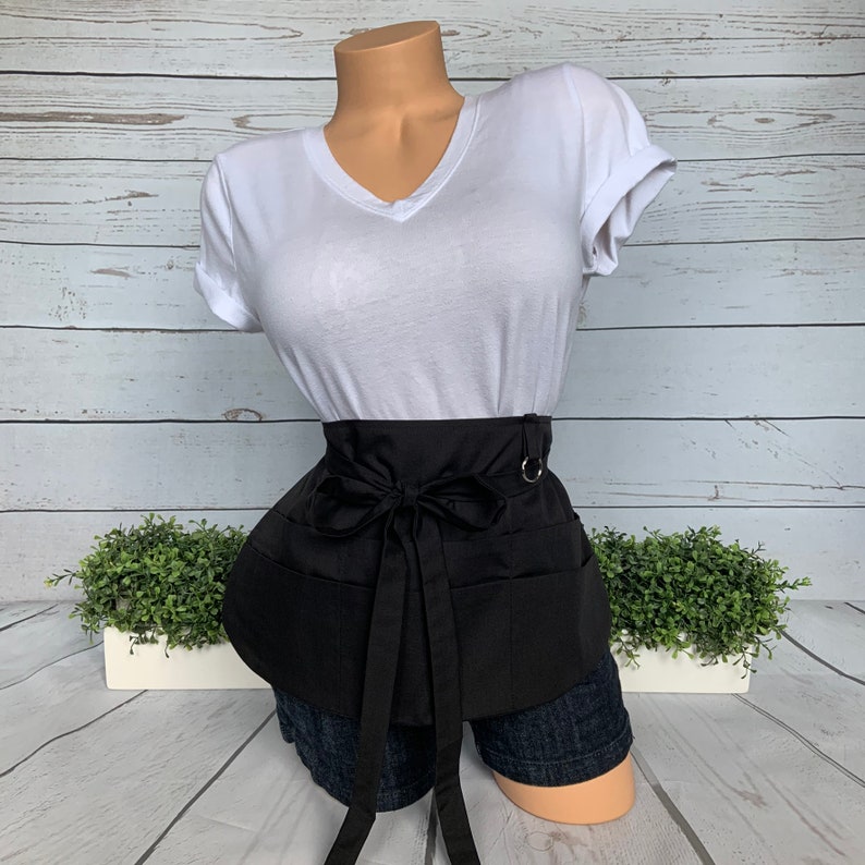 May include: Black apron with multiple pockets and a tie at the waist. The apron is worn over a white t-shirt and denim shorts.