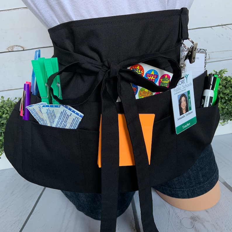 May include: Black teacher utility apron with multiple pockets, a large orange notepad, a name tag, and various school supplies. The name tag reads "Moore Public Schools" and "Amanda Terrance, Teacher".
