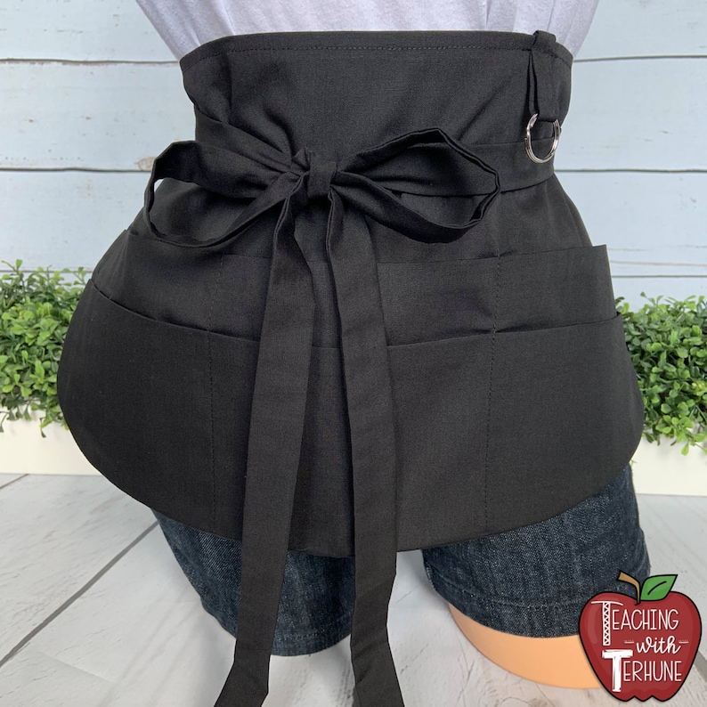 May include: Black apron with multiple pockets and a tie closure. The apron has a metal ring on the side.