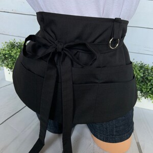 May include: Black apron with multiple pockets and a tie closure. The apron has a metal ring on the back.