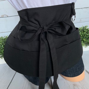 May include: Black apron with multiple pockets and a tie at the back. The apron has a silver ring on the side.