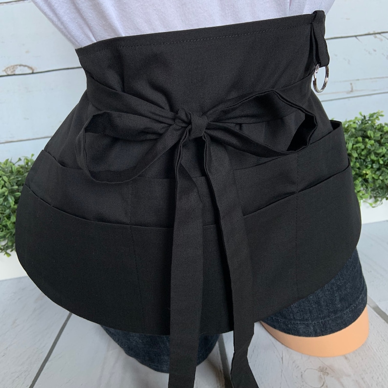 May include: Black apron with multiple pockets and a tie at the back. The apron has a silver ring on the side.
