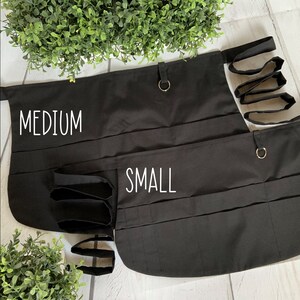 May include: Two black utility aprons with multiple pockets and loops. The larger apron is labeled "MEDIUM" and the smaller apron is labeled "SMALL".