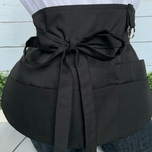 May include: Black utility apron with multiple pockets and a tie-style waist closure. The apron has a D-ring on the side.