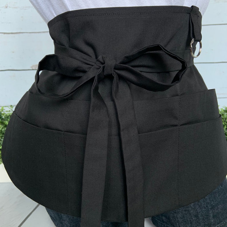 May include: Black utility apron with multiple pockets and a tie-style waist closure. The apron has a D-ring on the side.