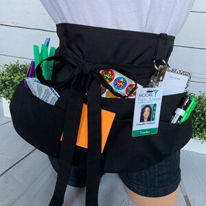May include: Black utility apron with multiple pockets, a black ribbon tie, and a green lanyard with a teacher's ID badge. The badge has a photo of a woman and the text "MOORE Public Schools LEARNING FOR LIFE W. Bonds Amanda Terbune Teacher".