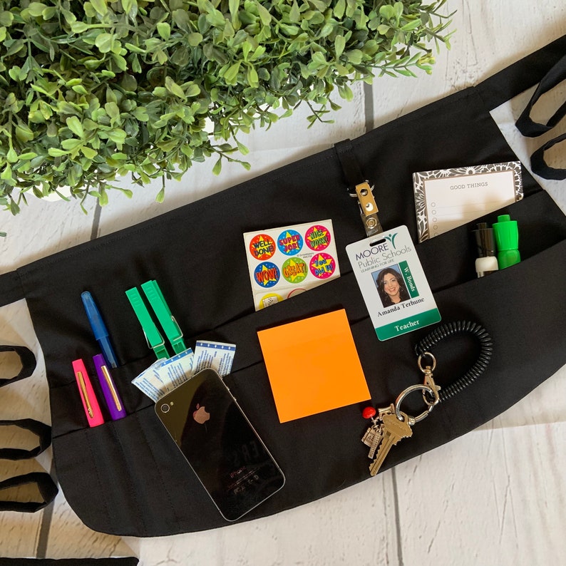 May include: Black utility apron with multiple pockets, featuring a cell phone, pens, stickers, a notepad, a key chain, and a teacher ID badge. The badge has a photo of a woman and the text "Moore Public Schools" and "Amanda Terhune, Teacher".
