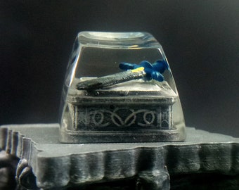 May include: A clear resin keycap with a miniature sword inside. The sword has a blue and yellow handle and a silver blade. The keycap is sitting on a gray pedestal.