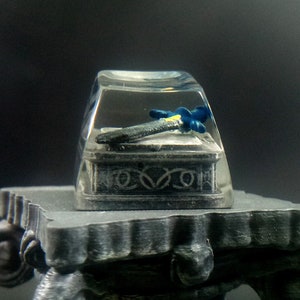 May include: A clear resin keycap with a miniature sword inside. The sword has a blue and yellow handle and a silver blade. The keycap is sitting on a gray pedestal.