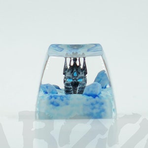 May include: A clear resin keycap featuring a blue and white icy landscape with a black and silver miniature castle with blue glowing windows.