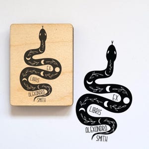 Può includere: Un timbro in legno rettangolare con un'illustrazione di un serpente nero con fasi lunari e motivi a foglie, accanto a un'impronta a inchiostro nero su carta bianca. Il design include il testo "EX LIBRIS ALEXANDRA SMITH" nel corpo del serpente.