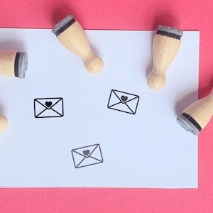 Pode incluir: Quatro carimbos de madeira com almofadas de tinta pretas e um design simples de um envelope com um coração dentro. Os carimbos estão sobre uma superfície branca.