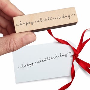 May include: A wooden stamp with the words "happy valentine's day" in a cursive font is held above a white tag with the same text. A red ribbon is tied to the tag, creating a festive look.
