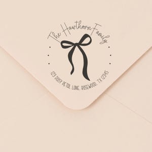 Op de afbeelding: Een crèmekleurige envelop met een cirkelvormige stempel met de tekst "The Hawthorn Family" en adresgegevens. De stempel bevat een zwart strikdesign in het midden. Het adres luidt "123 Daisy Petal Lane, Rosewood, TX 12345".