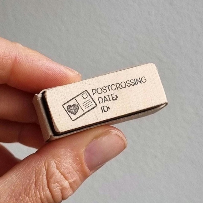May include: A rectangular wooden rubber stamp featuring a laser-engraved design of a postcard with a heart icon. The text on the stamp reads POSTCROSSING, DATE:, and ID:. The stamp has a light-coloured wooden block base and a black rubber surface.