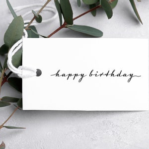 May include: A rectangular white paper gift tag with a hole reinforced by a dark metal eyelet and threaded with white string. The text "happy birthday" is printed in a black, thin, cursive script font across the center of the tag.