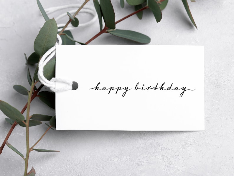 May include: A rectangular white paper gift tag with a hole reinforced by a dark metal eyelet and threaded with white string. The text "happy birthday" is printed in a black, thin, cursive script font across the center of the tag.