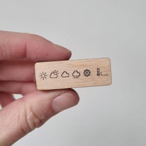 Può includere: Un piccolo blocco di legno rettangolare con simboli meteorologici incisi. I simboli includono un sole, nuvole, pioggia, fiocchi di neve e un termometro. Il blocco è tenuto nella mano di una persona.