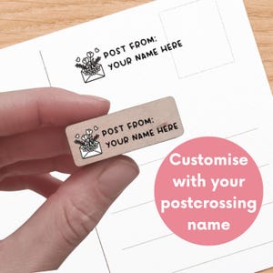 Könnte beinhalten: Ein rechteckiger Holzstempel mit einem schwarzen Motiv eines Briefumschlags, aus dem Blumen und Herzen ragen, sowie dem Text "POST FROM: YOUR NAME HERE". Der Stempel ist vor dem Hintergrund einer Postkartenvorlage abgebildet.