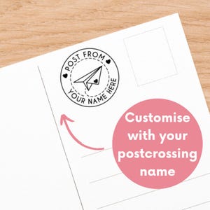 Peut inclure: Carte postale blanche avec un tampon circulaire noir représentant un avion en papier et le texte "Post From Your Name Here". Un cercle rose avec le texte "Customise with your postcrossing name" est également présent.