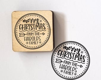 May include: A square wooden rubber stamp and its circular black ink impression. The design features a circular border with stars, the text "merry CHRISTMAS", an arrow graphic, and the text "FROM THE HAROLDS FAMILY" with snowflake accents.