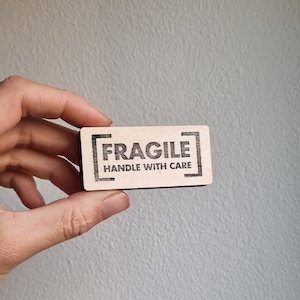 Könnte beinhalten: Ein Holzschild mit dem Text "[FRAGILE] HANDLE WITH CARE" in schwarzen Buchstaben.