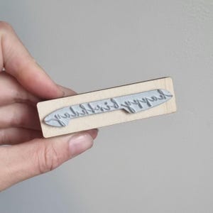 May include: A rectangular wooden stamp featuring a raised gray rubber surface with the text "happy birthday" written in a cursive script. The stamp is designed for manual application and displays the text in a mirrored orientation for printing.