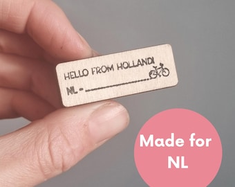 May include: Wooden stamp with the text "HELLO FROM HOLLAND" and a bicycle icon. The stamp also has the text "NL" and a blank line for personalization. Made for NL.