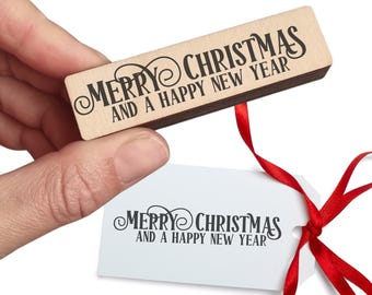 May include: A wooden stamp with the words "MERRY CHRISTMAS AND A HAPPY NEW YEAR" in black script. A white gift tag with the same text is attached with a red ribbon. The stamp is held by a hand.