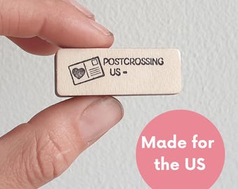 May include: A wooden stamp with the text "Postcrossing US -" and a small image of a postcard with a heart. The stamp is made for the US.