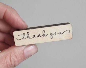 May include: A rectangular light-colored wooden block featuring the words thank you written in a black cursive script. The block has a smooth finish and a dark brown edge, presenting a simple and clear message on a natural wood surface.