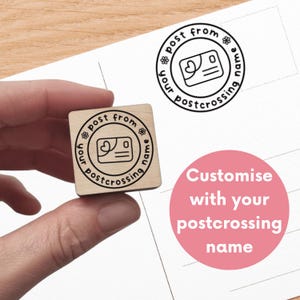 Op de afbeelding: Een vierkante houten rubberstempel met een cirkelvormig ontwerp met de tekst post from your postcrossing name en een klein ansichtkaartpictogram met een hartje. De stempel maakt een zwarte inkt-afdruk op het papier.