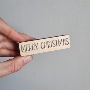 Op de afbeelding: Een rechthoekig houten bord met de woorden "MERRY CHRISTMAS" in een handgeschreven lettertype. Het bord is lichtbruin met donkerbruine tekst. Het bord wordt in de hand van een persoon gehouden.