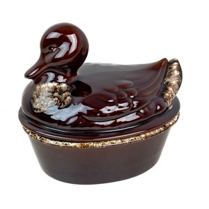 May include: A brown ceramic casserole dish shaped like a duck. The dish has a glazed finish and a brown and white rim.